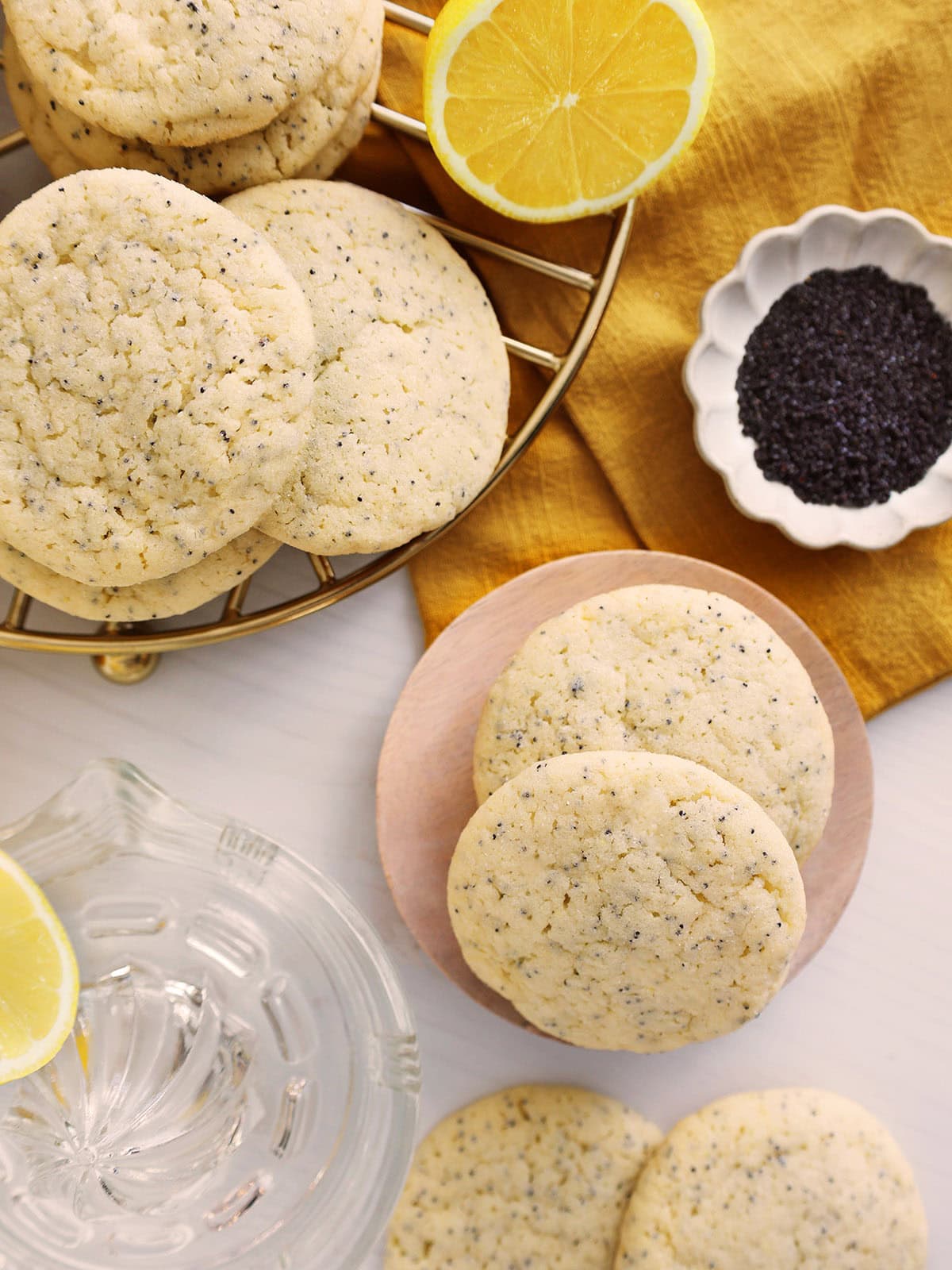 Lemon poppyseed cookies scattered across a wire rack, small plate and a countertop next to poppyseeds and lemons.