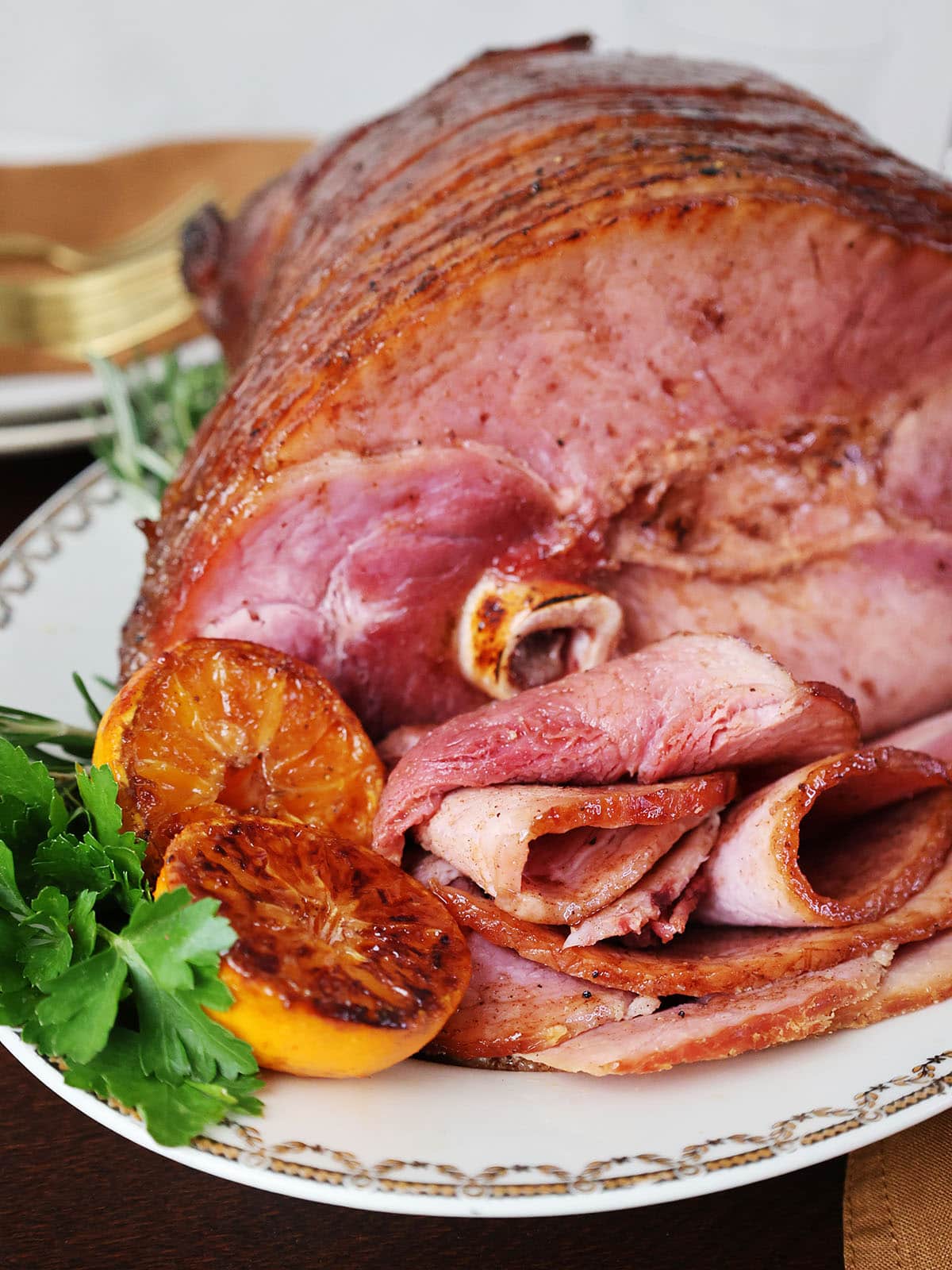Close up of a glazed ham on a white platter next to fruit and greens.