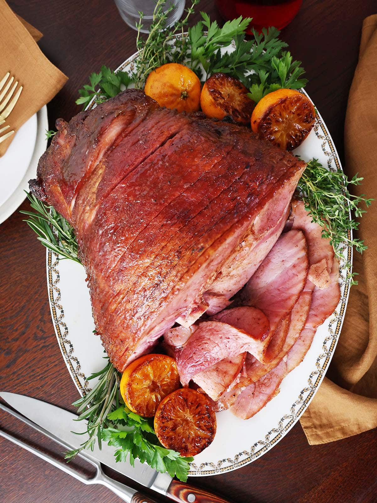 Top view of a glazed roasted ham on a white platter next to greens and fruit.