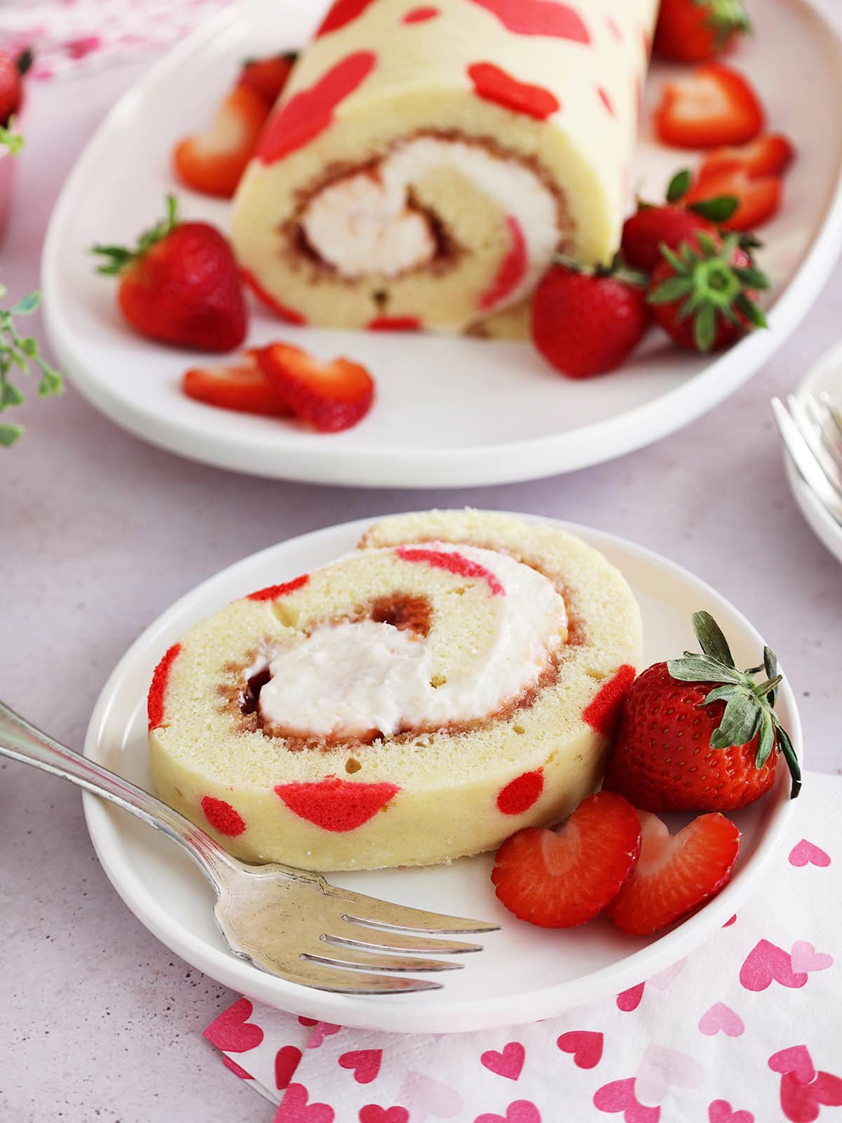 A slice of Valentine's Day strawberry cake roll on a small white plate next to strawberries with the rest of the cake in the background.