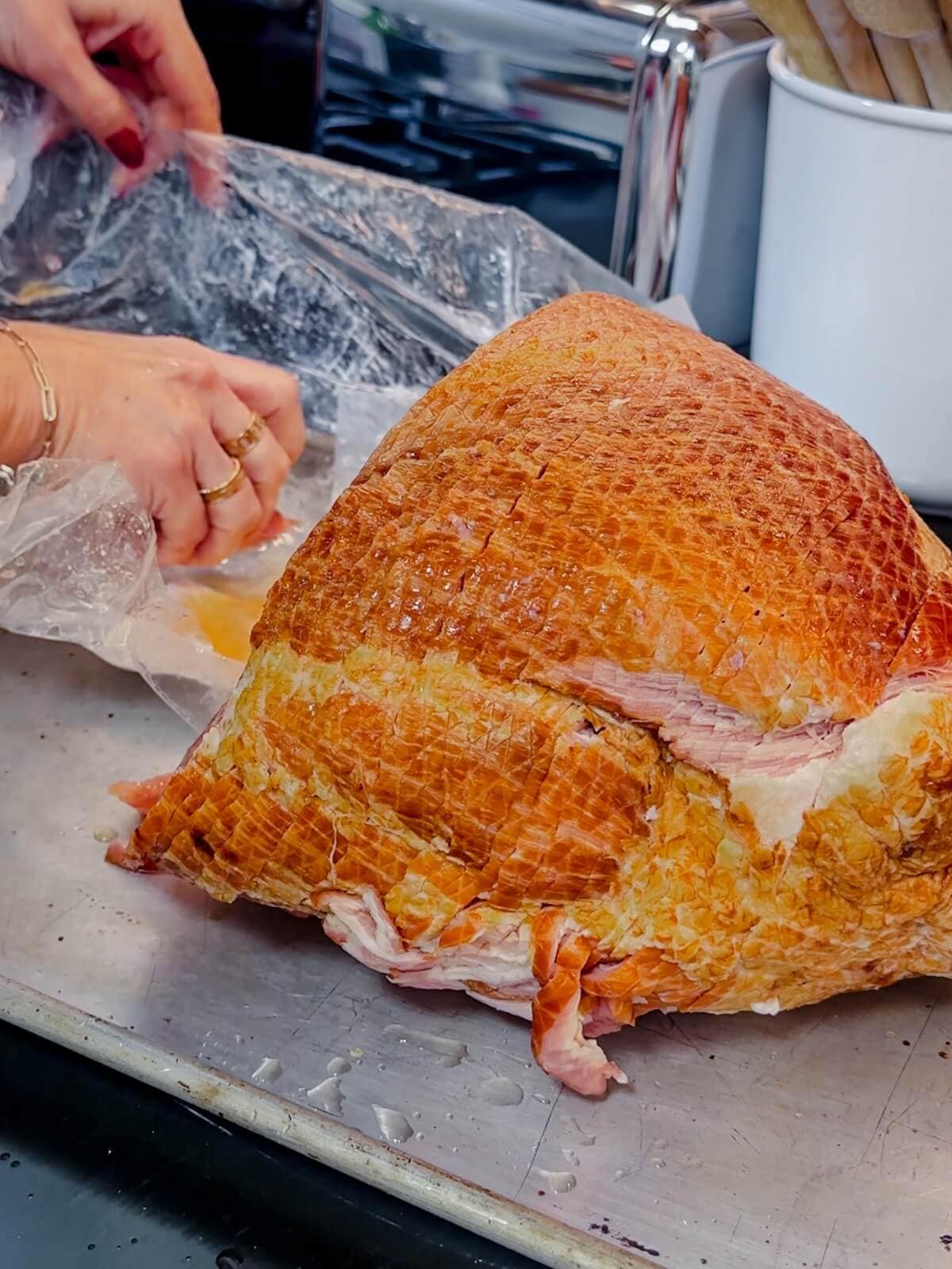 Hand sliding the wrapper away from a ham on a baking tray.
