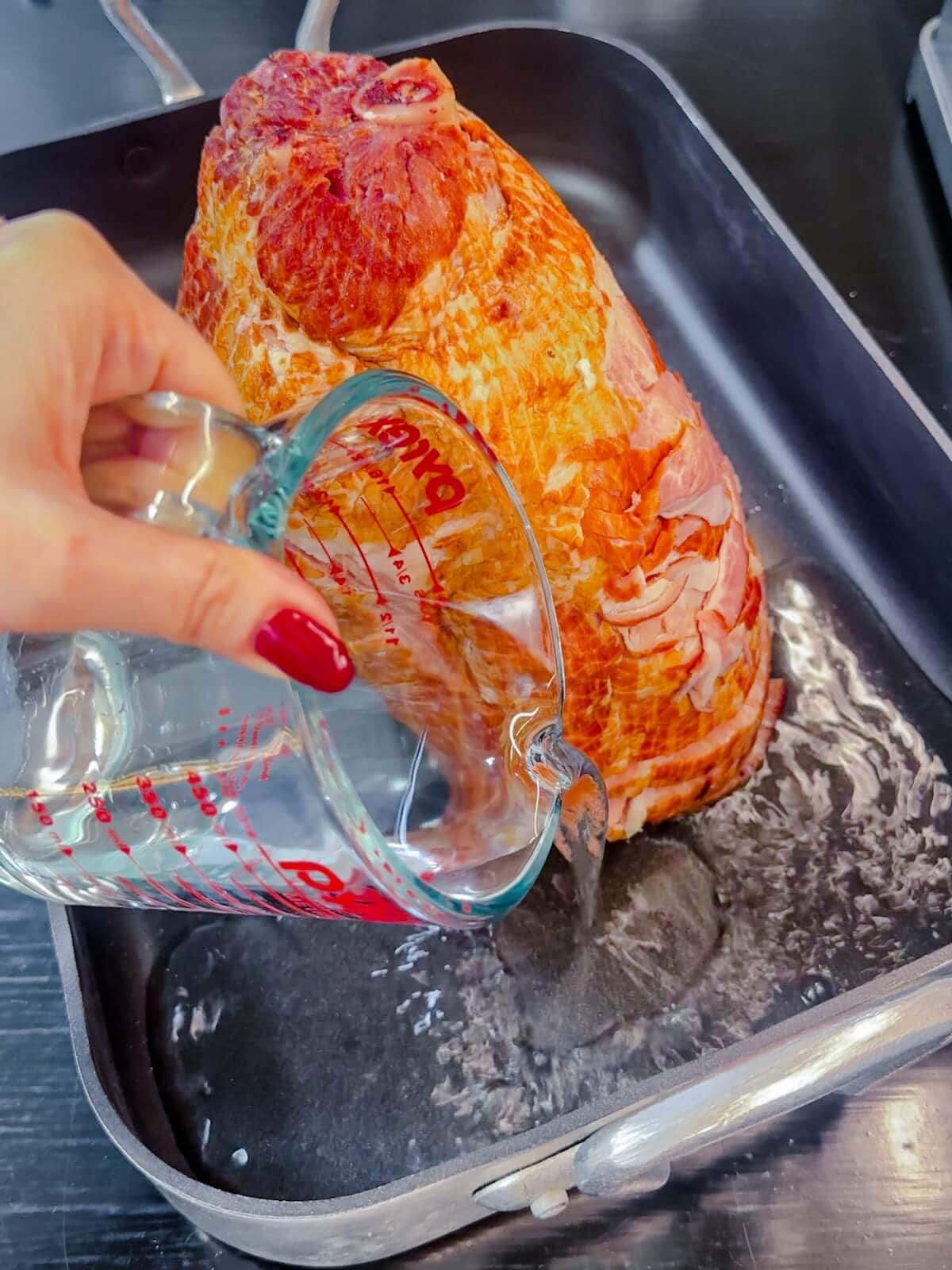 Hand pouring water into the bottom of a roasting pan that already has a ham in it.