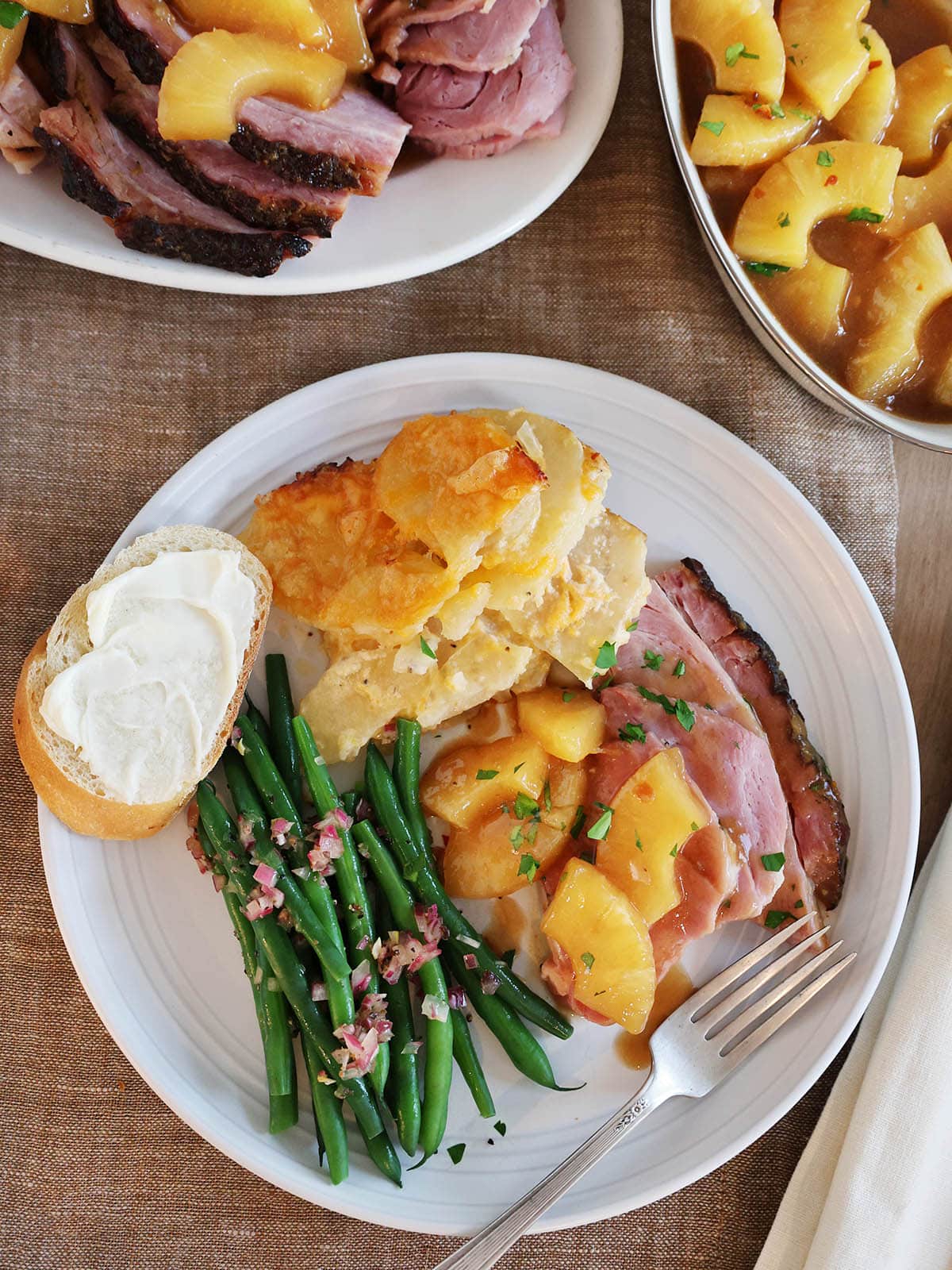 Overhead view of a dinner plate with a piece of bread, potatoes, green beans, and ham topped with pineapple.