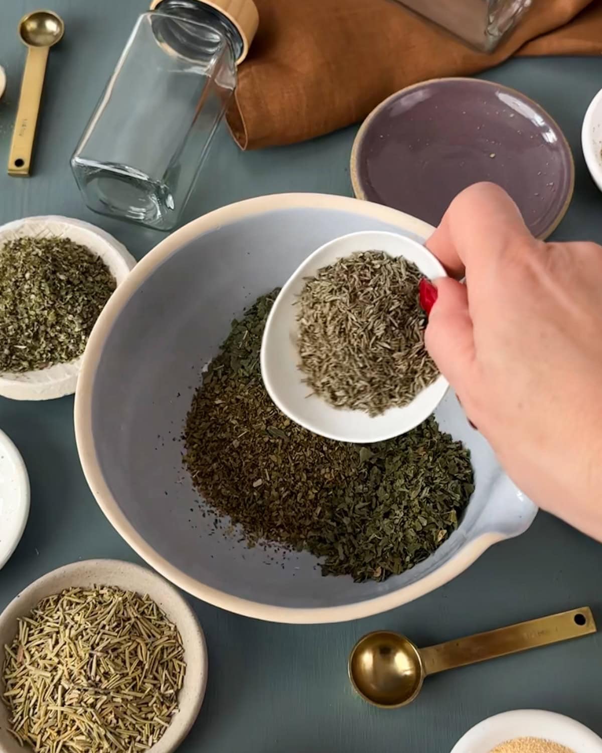 Adding measured dried herbs to a bowl to make Italian seasoning.