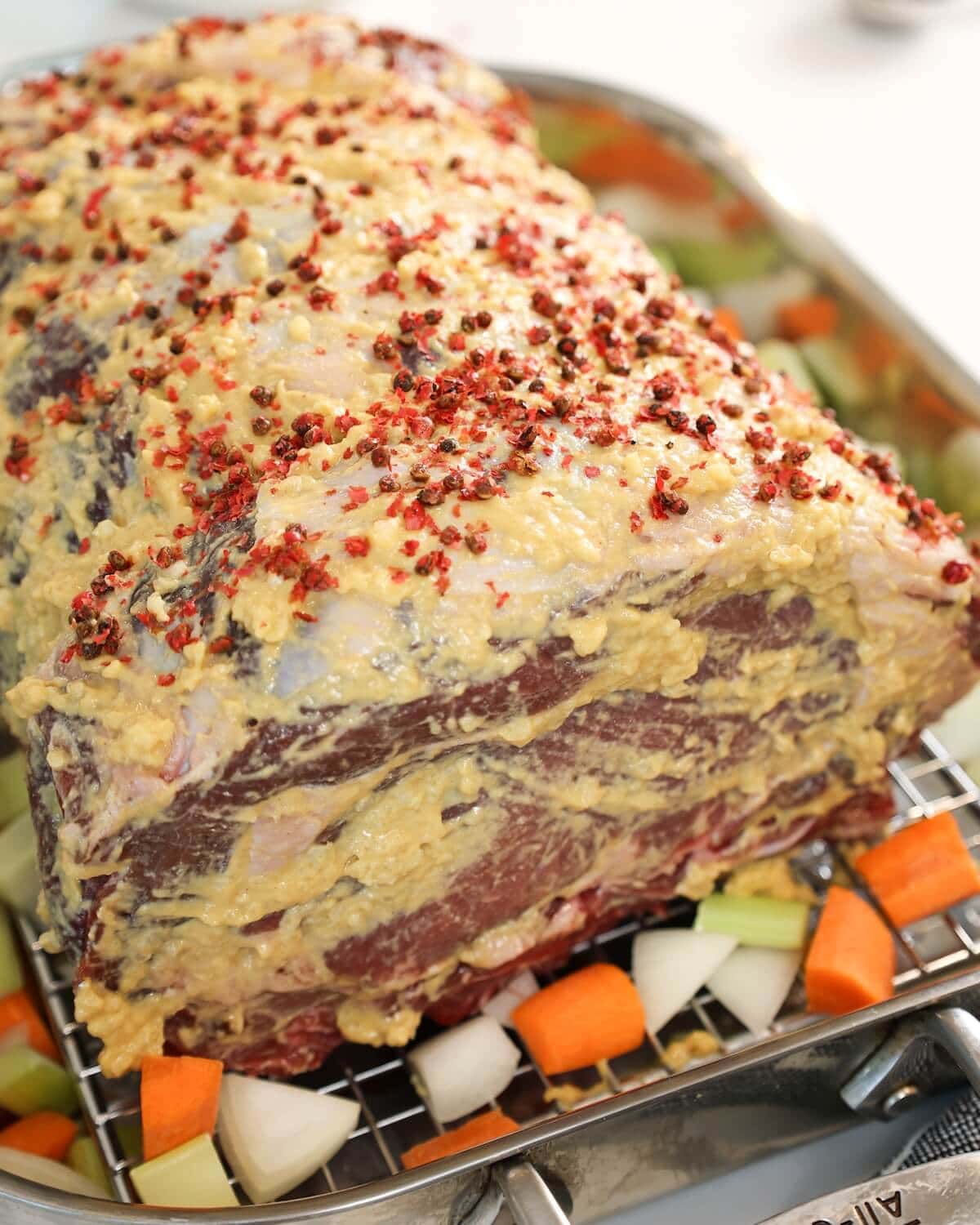 Prime rib roast in a roasting pan with mustard rub and chopped carrots and onions around the base.