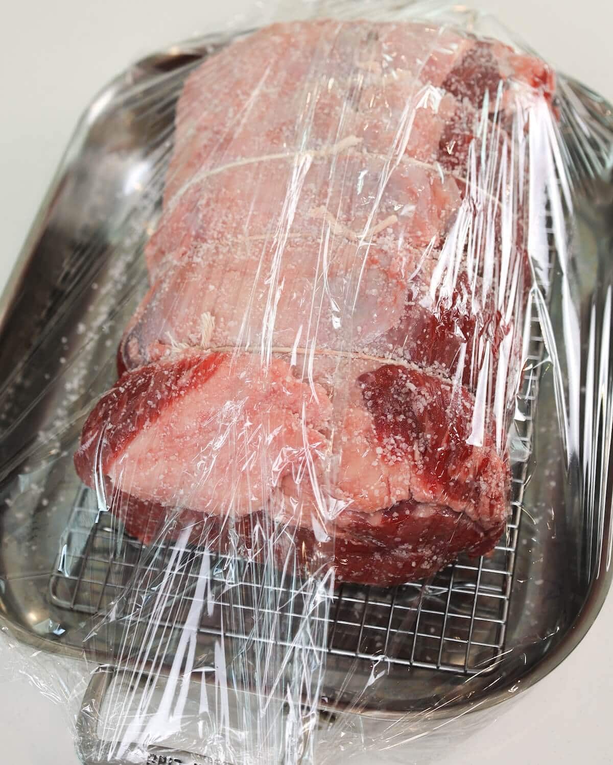 A large prime rib roast rubbed with salt in a roasting pan completely wrapped in plastic wrap.
