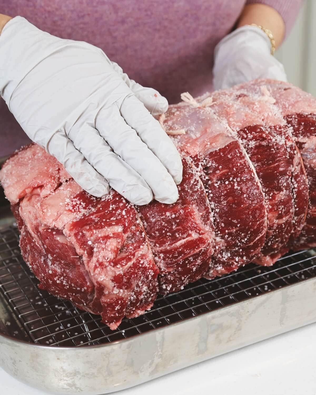 Hands with plastic gloves rubbing kosher salt on a prime rib roast in a roasting pan.