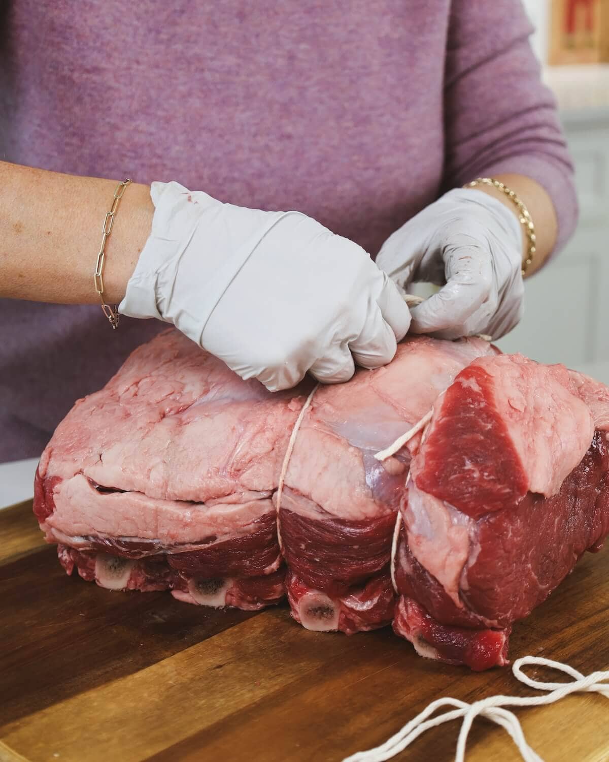 Hands in plastic gloves tying kitchen twine on a large, prime rib roast to keep rib bones and meat together for roasting.
