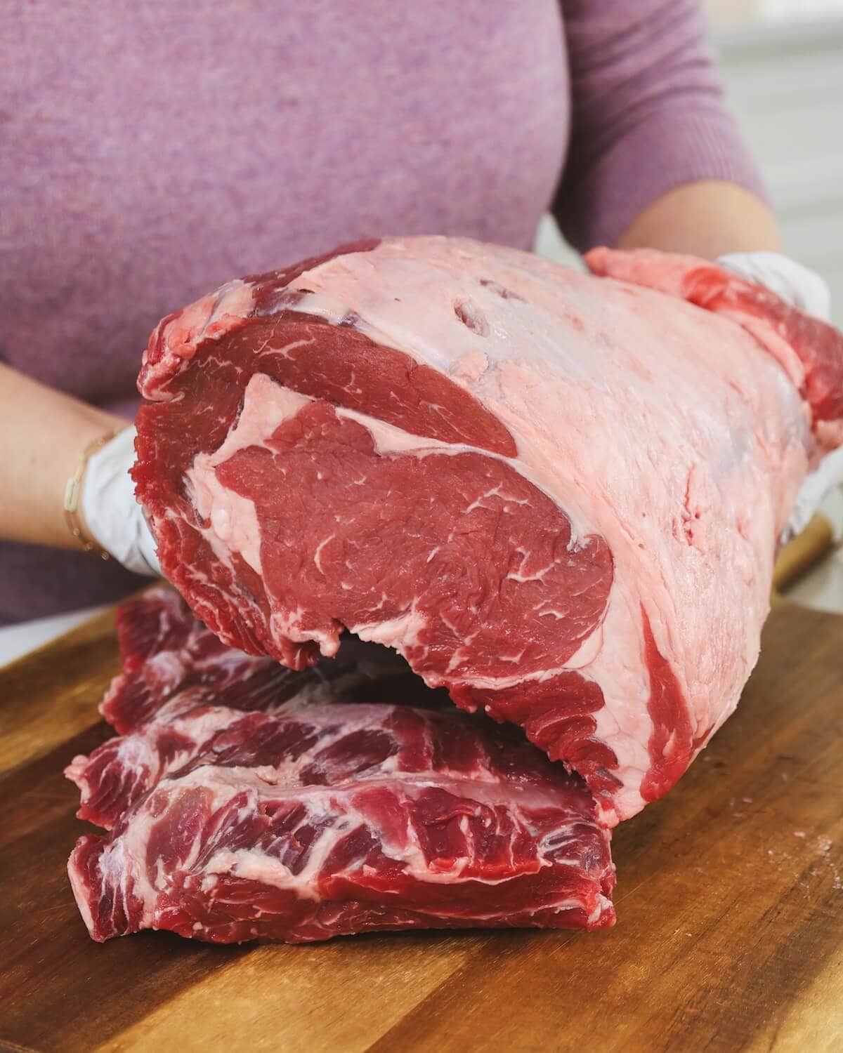 Hands in plastic gloves placing a prime rib roast on top of rib bones on a cutting board.