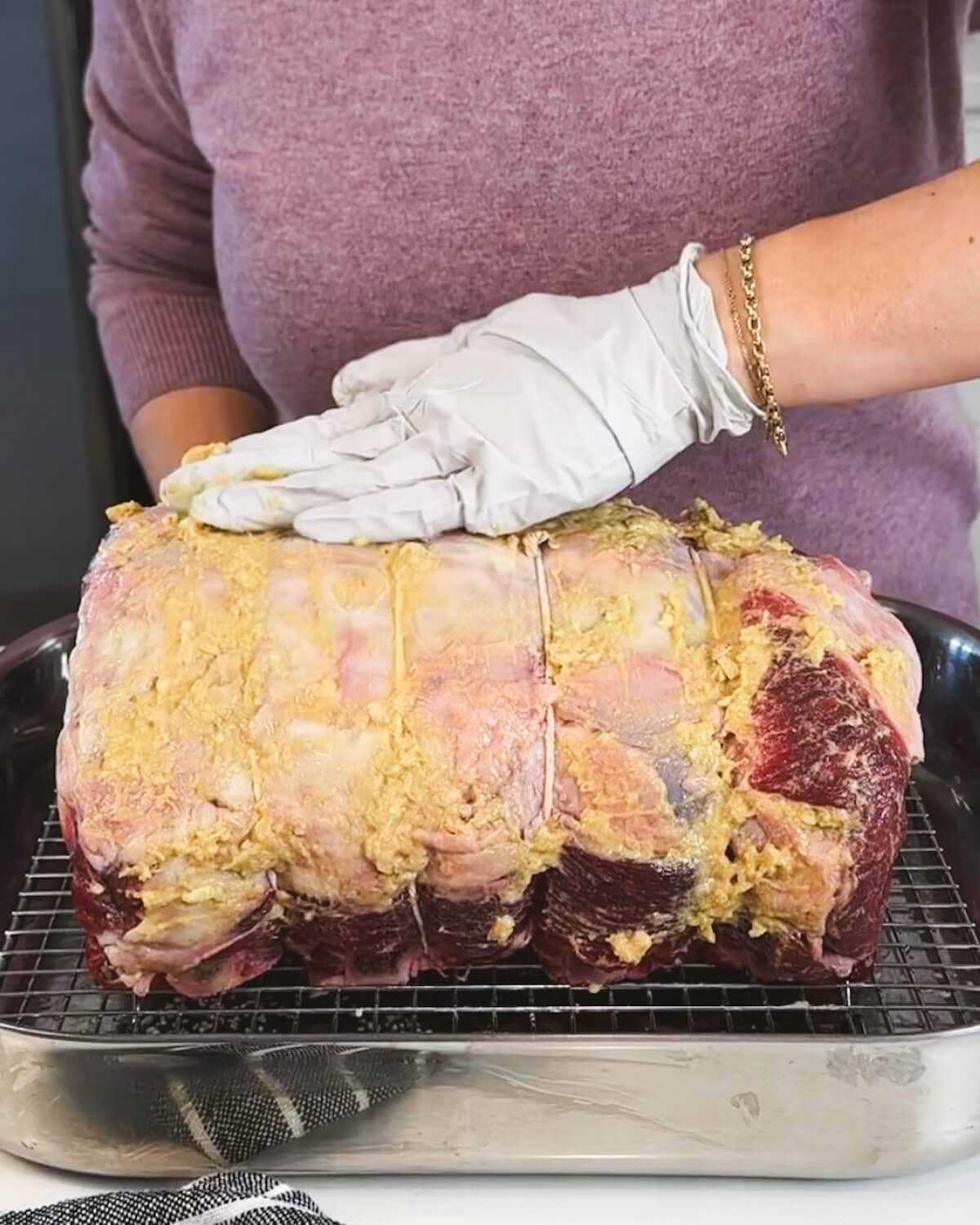 Hands wearing gloves applying a thick mustard and garlic paste to the surface of a raw beef roast.