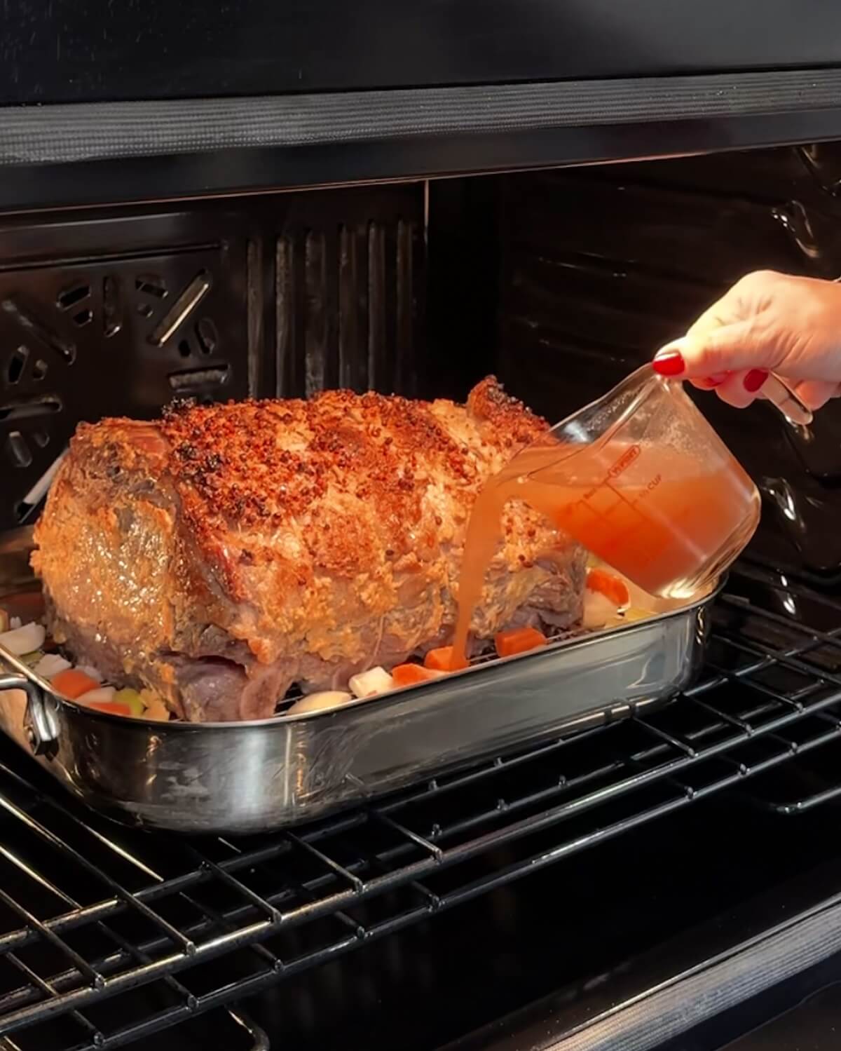 A prime rib roast in a roasting pan in the oven partially cooked with hands pouring broth into the pan.