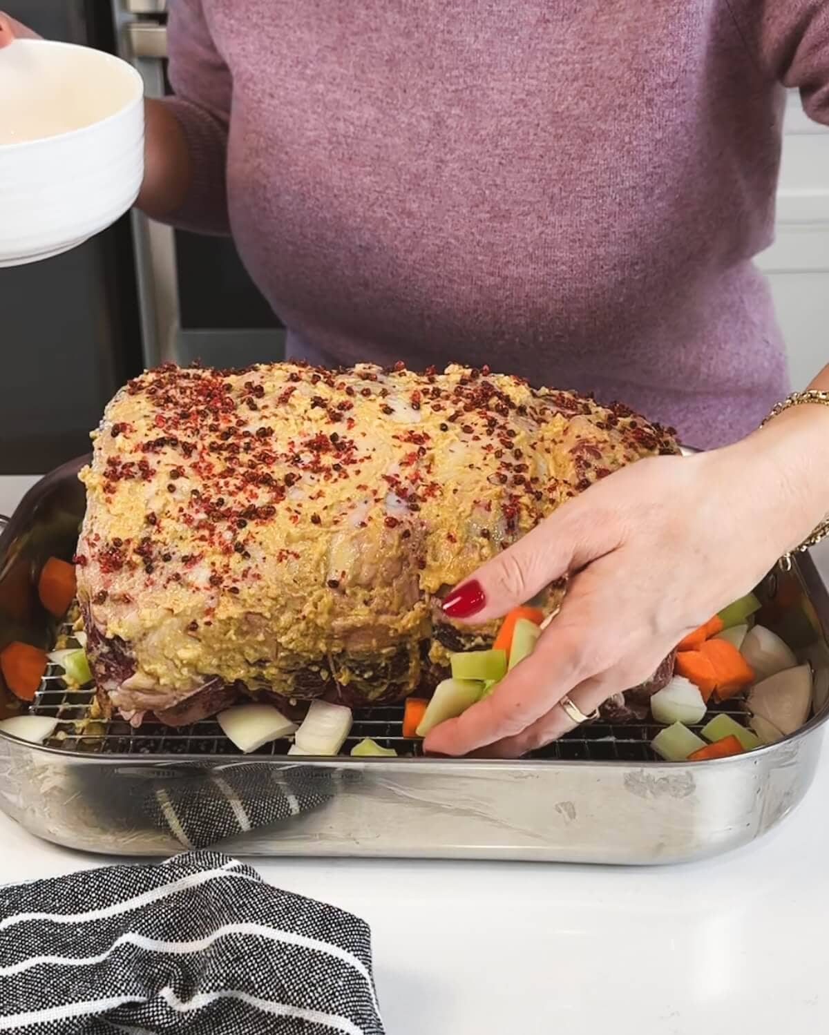 A close-up of a seasoned prime rib roast in a roasting pan with chopped carrots and onions around the base.