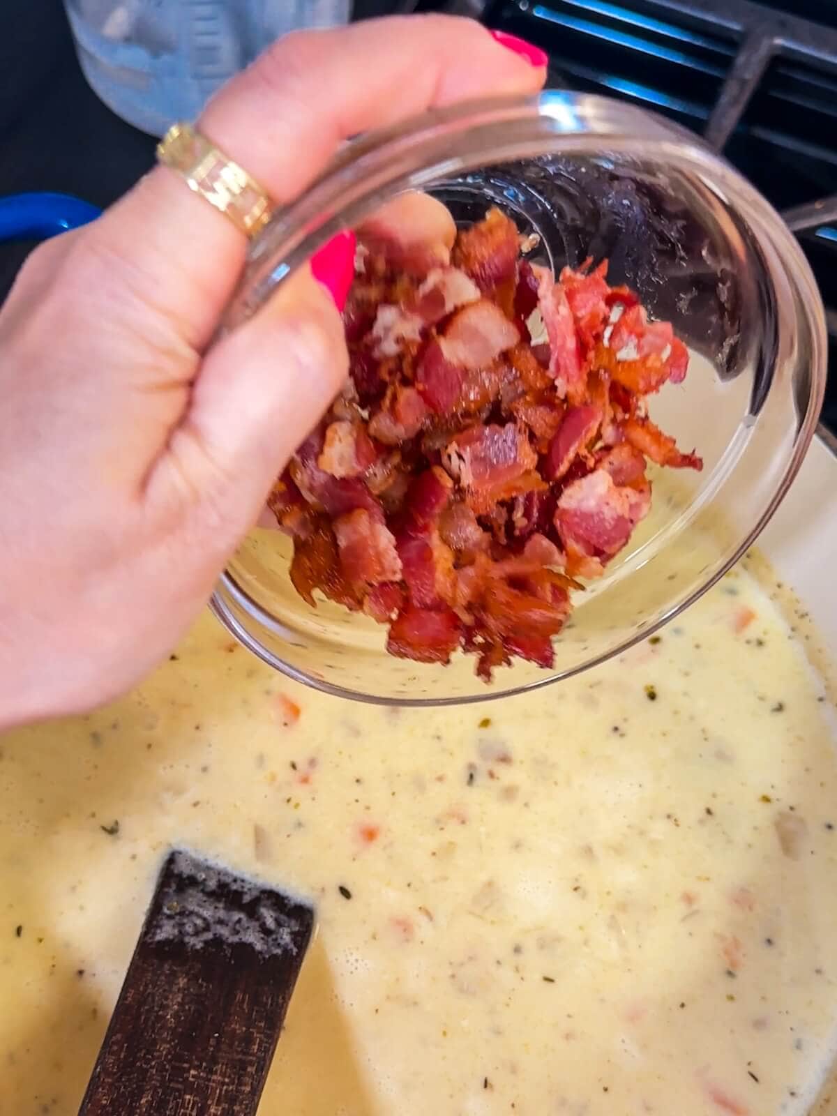 Hand pouring in crispy bacon into a pot of potato soup.