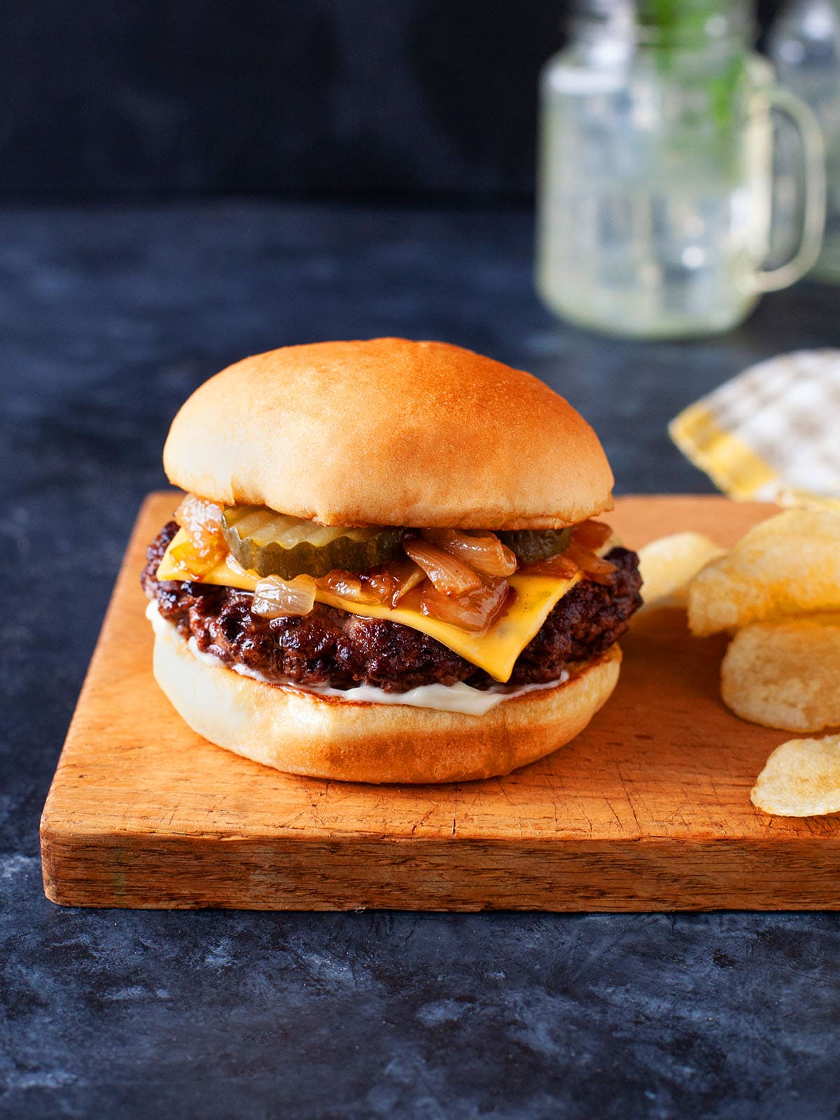 A butter burger on a wooden cutting board next to chips. 