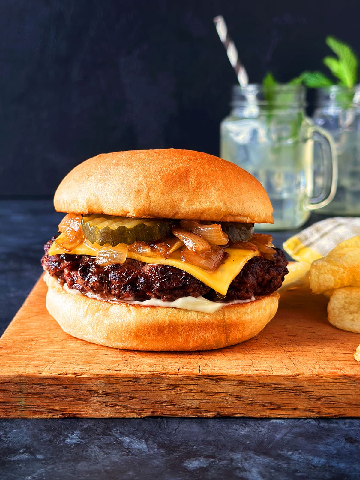 Close up of a butter burger on a wooden cutting board next to chips and mason jar drinks.
