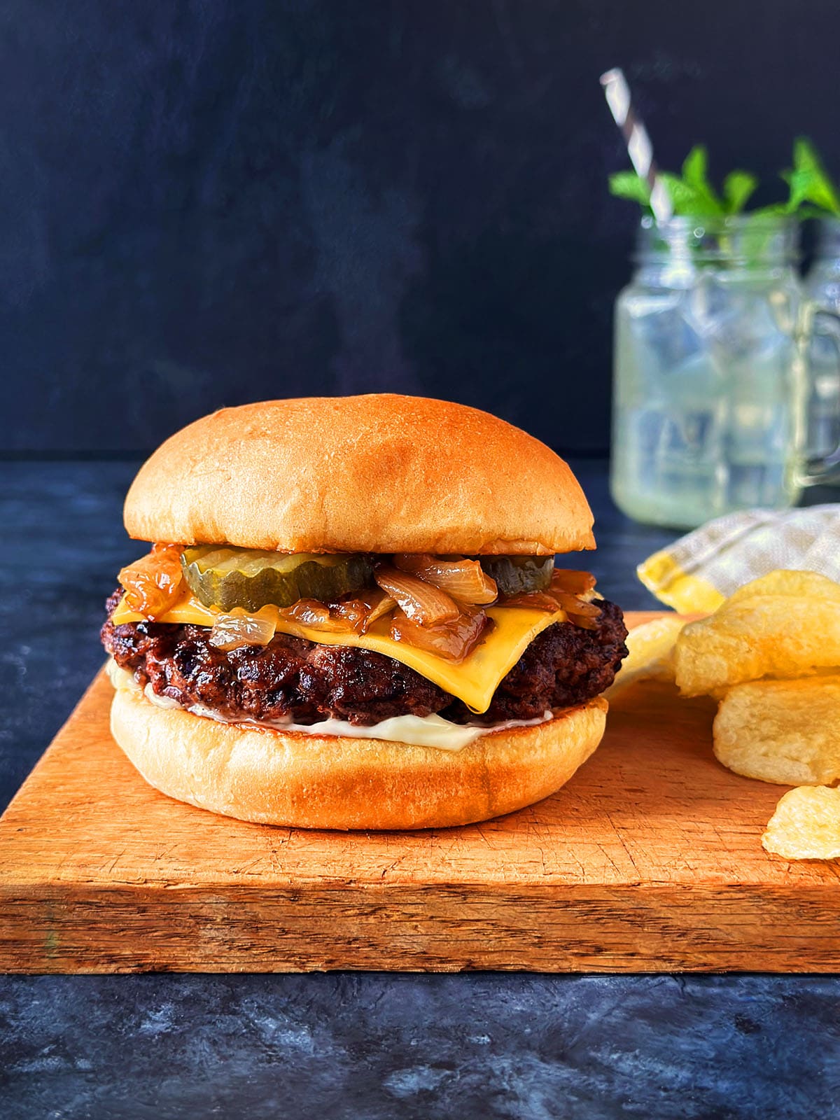 A butter burger on a wooden cutting board next to chips and a mason jar drink. 