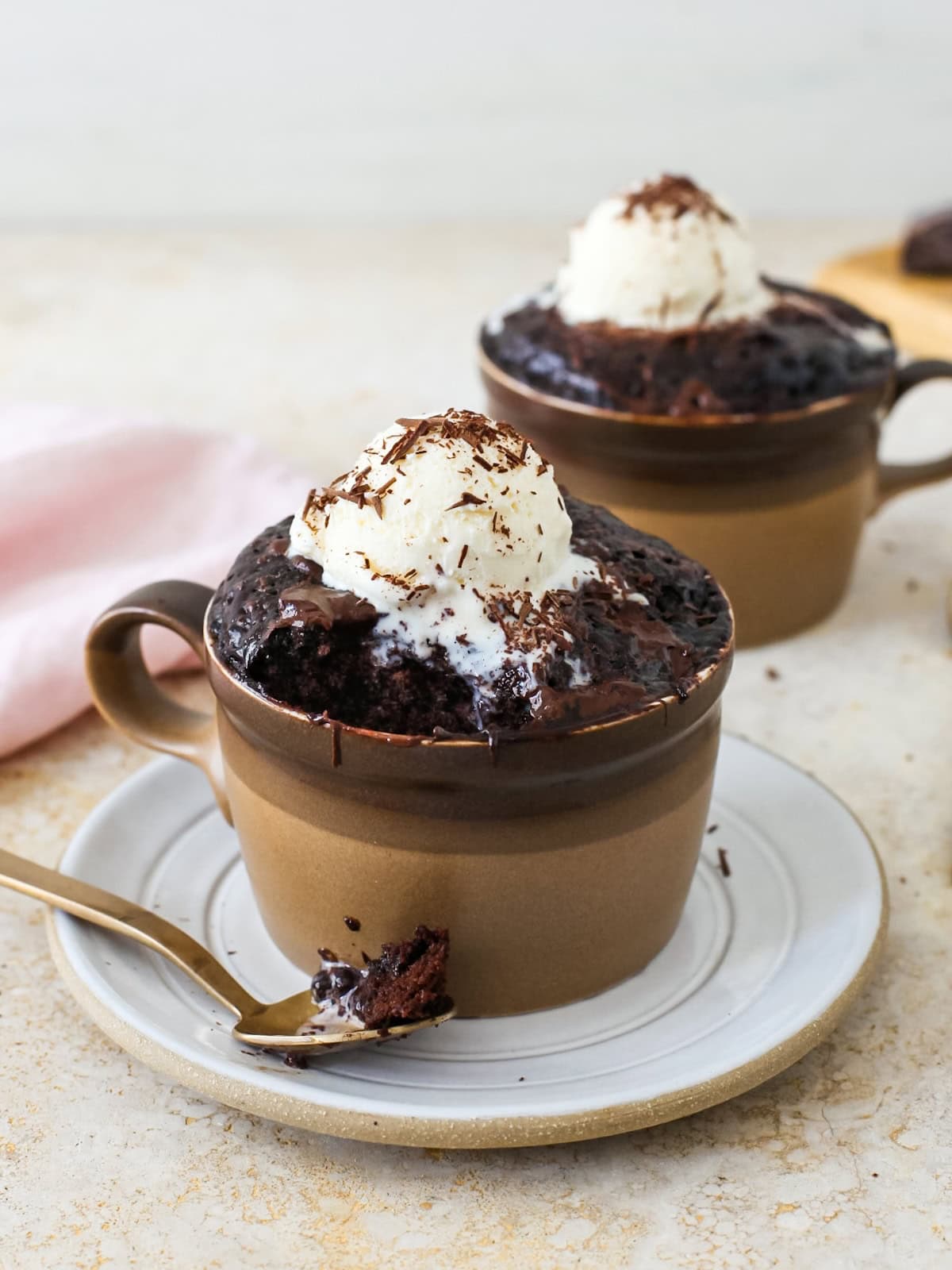 Two chocolate mug cakes with a bite removed from the front cup on a spoon.