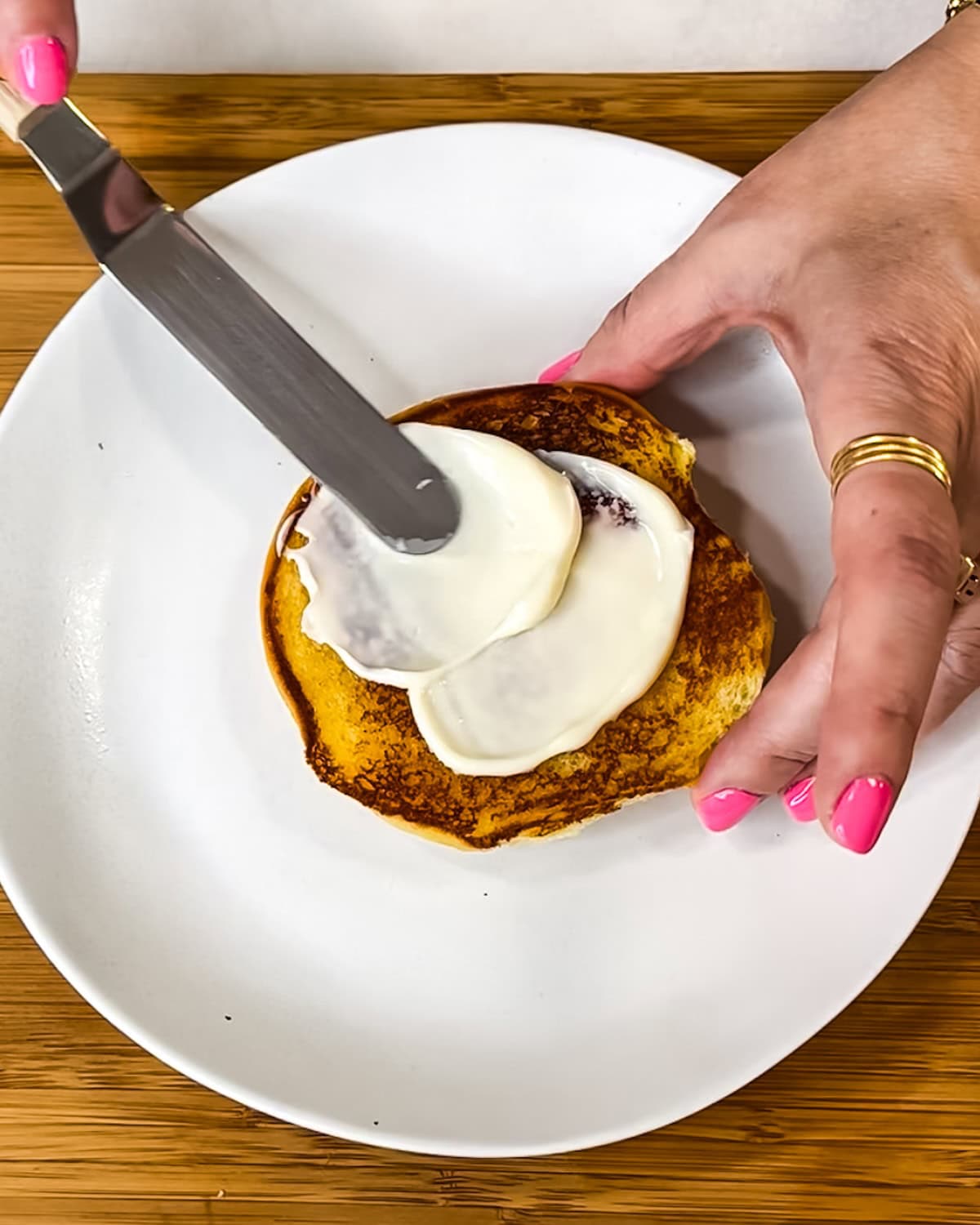 Hand spreading mayo on top of a burger bun.