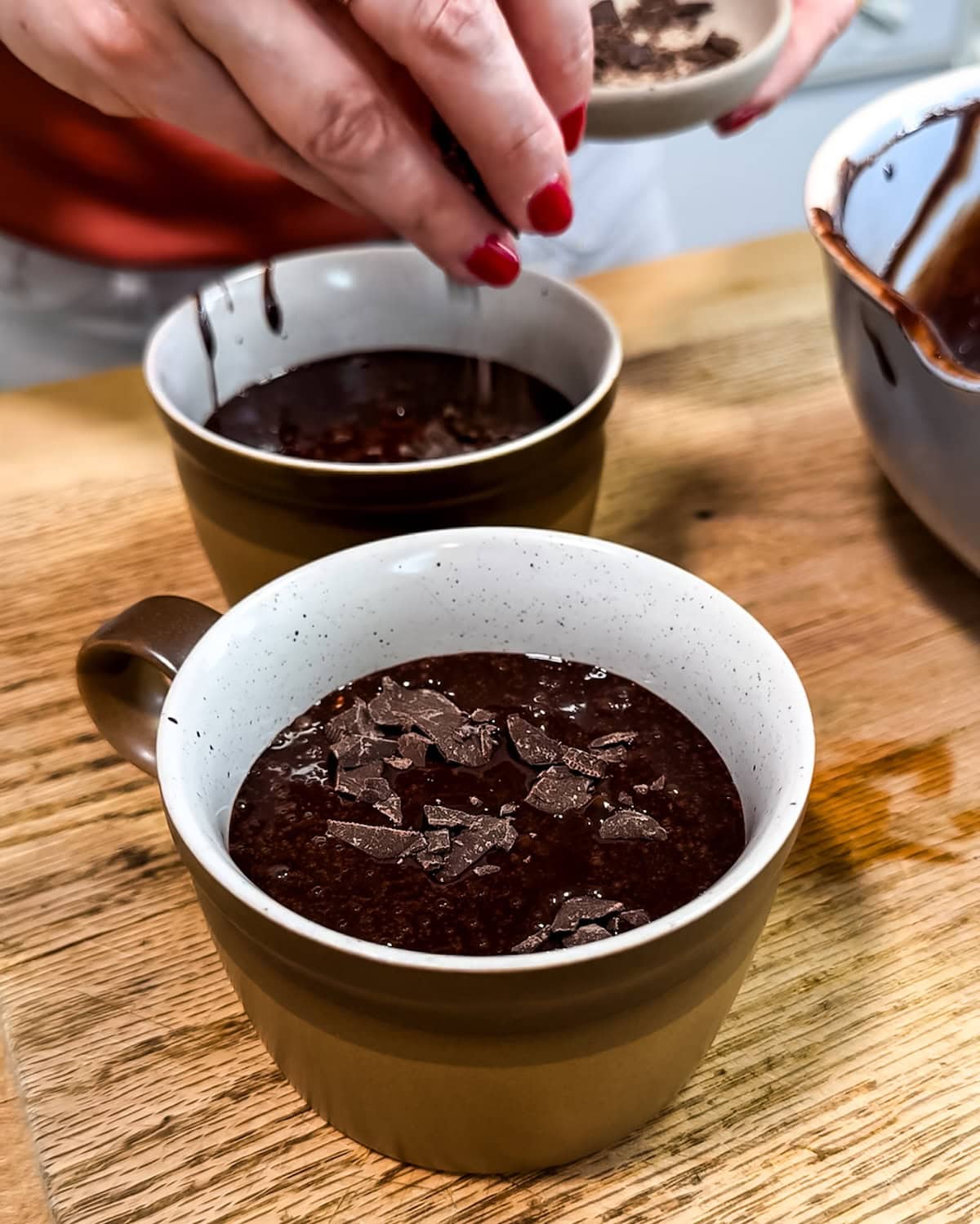 Two mugs with cake batter being sprinkled with chocolate chunks. 