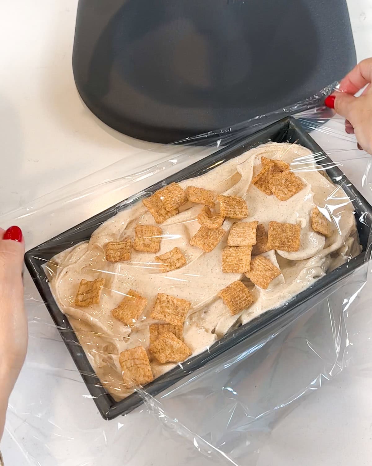 Spreading plastic wrap over the top of a pan of cinnamon toast crunch ice cream.