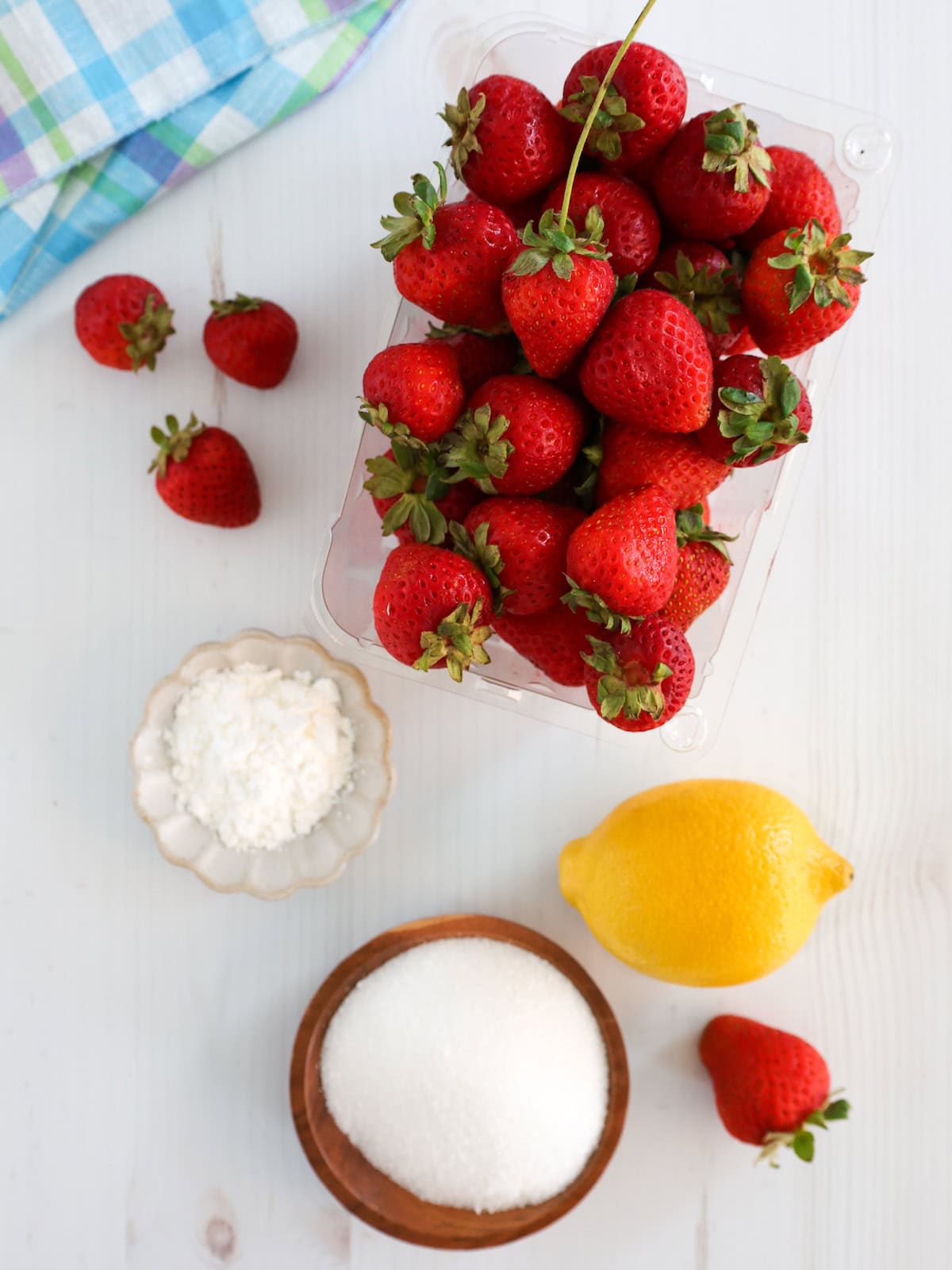 ingredients for strawberry sauce on a white table including lemon, strawberries, sugar and cornstarch.