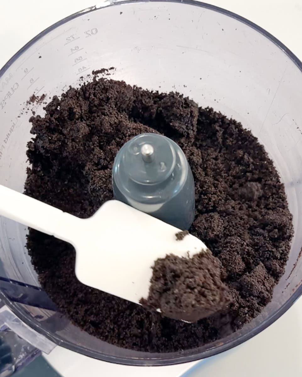 Spatula holding up finely ground Oreo cookies over a food processor.