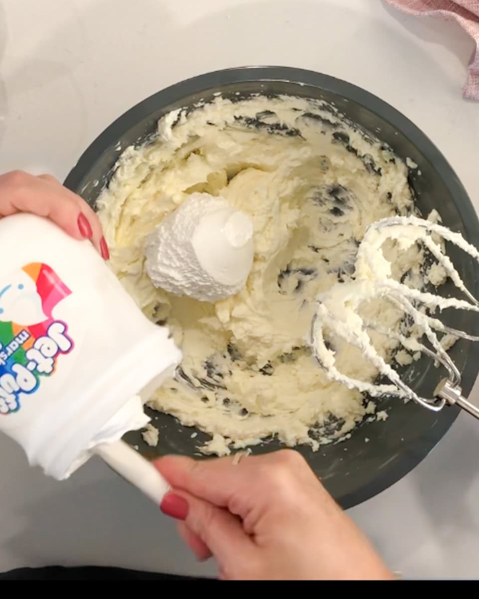A mixing bowl with whipped cream cheese and a hand adding marshmallow creme.