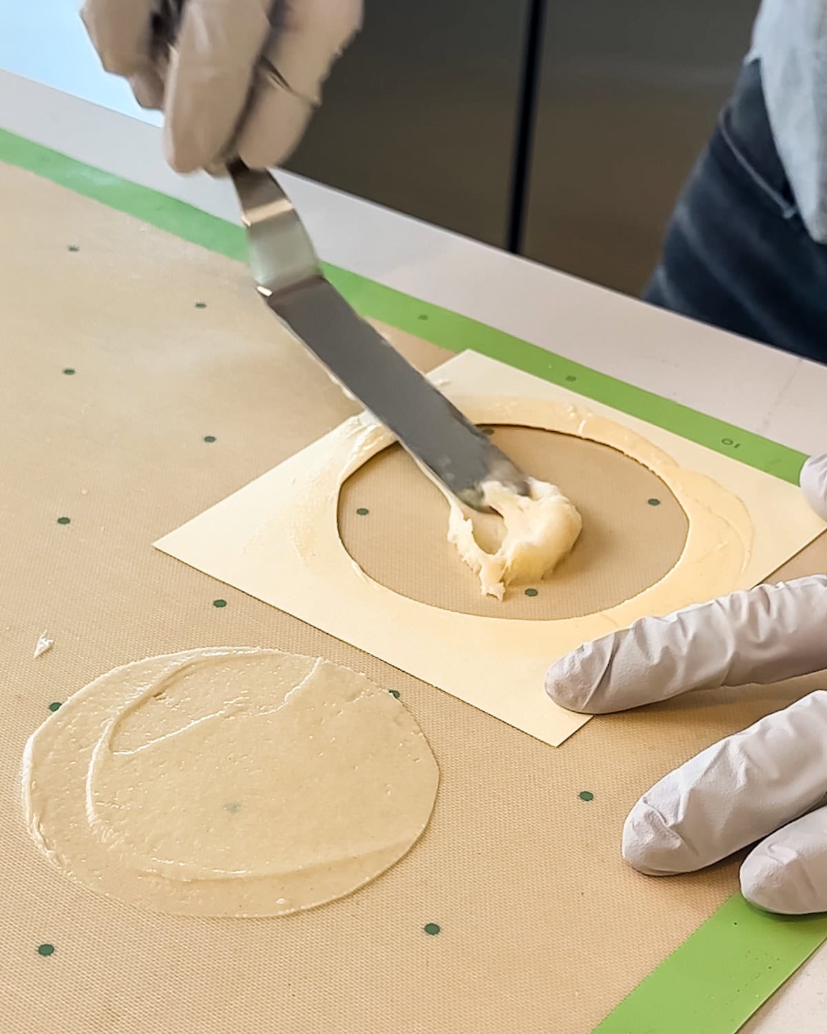 Spreading pirouette cookie batter into a template next to another formed cookie on a silicone baking mat.